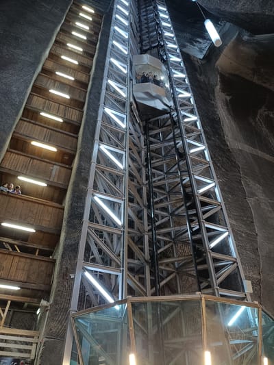 The image shows a large, open shaft in a salt mine, illuminated by numerous fluorescent lights. A modern elevator with a glass enclosure is ascending the shaft, carrying several people. To the left of the elevator shaft, a wooden staircase with attached lights runs along the rough, textured walls of the mine. The salt mine is a significant tourist attraction in Turda, Romania.