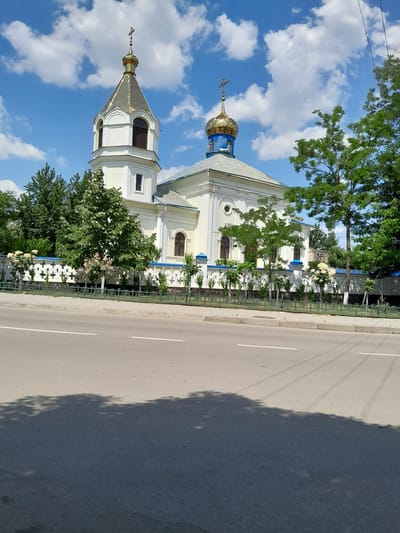 The image shows a white church with a gold dome and a tall bell tower. The church is located on a street in the city of Cimișlia, Moldova. There are trees and a white fence in front of the church. The street is asphalt with white lines. The sky is blue with white clouds. The image is taken from a low angle, looking up at the church.