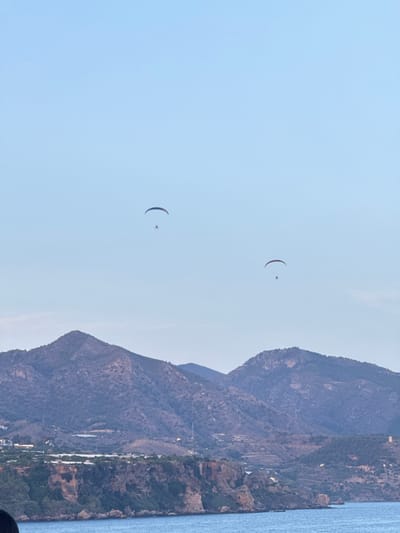 Two paragliders are visible in the sky over the coastal city of Nerja, Spain. The paragliders are at different altitudes and distances from the viewer. The foreground of the image features the Mediterranean Sea, with a rocky coastline and cliffs rising in the middle ground. Beyond the coastline, a mountainous landscape with scattered vegetation and some buildings is visible. The sky is clear and blue, indicating daytime.