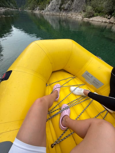 The image shows a first-person perspective from inside a yellow raft on a river. The raft is textured with water droplets, suggesting recent exposure to water. In the foreground, a person's legs are visible, wearing pink Crocs on one foot and white sneakers on the other. The person is wearing white shorts on one leg and black leggings on the other. The river water is clear and a vibrant turquoise color, with rocky banks and green foliage visible in the background. The overall scene depicts an outdoor adventure activity, likely rafting, on a scenic river. There is no specific indication of the location being in Džajići, Bosnia and Herzegovina.
