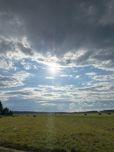 The image depicts a wide, grassy field dotted with numerous round hay bales. The field stretches towards a treeline in the distance, behind which are visible some power lines and scattered buildings. The sky above is dramatic, with a mix of dark, heavy clouds and bright, sunlit patches. The sun itself is visible, casting a bright ray of light through the clouds. The environment appears to be rural and agricultural, possibly located in the vicinity of Lukovnya, Russia, given the agricultural landscape and the style of the hay bales. There are no people or animals visible in the frame. The overall impression is one of a vast open space under a dynamic, weather-beaten sky.