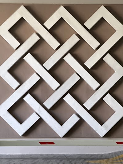This media file features a close-up, overhead shot of a white geometric pattern against a brown background. The pattern is composed of interconnected, L-shaped elements that create a sense of depth and complexity. The lighting suggests an indoor setting, possibly a wall or decorative panel, with subtle shadows cast by the raised elements. There are no people visible, and the focus is entirely on the abstract design.

On the right side of the image, a vertical strip of lighter material is visible, bordered by a thin green line. A small red label with white text, reading "BFC", is affixed to this strip. Beyond this, another textured surface with faint grey and yellow lines can be seen, possibly indicating an adjacent wall or architectural feature.

The overall impression is one of clean lines and modern design. The lack of specific environmental cues makes it difficult to pinpoint the exact location beyond the provided context of Santa Marta, Colombia. There is no discernible activity or event taking place. The mood is neutral and purely descriptive, focusing on the visual elements of the pattern and its immediate surroundings.
