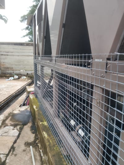 This image depicts the exterior of a large industrial cooling unit, likely an air conditioning or refrigeration system, situated outdoors. The unit is encased in a brown metal structure with visible ventilation fins and a protective wire mesh cage surrounding it. The scene appears to be in a somewhat neglected urban or industrial setting, with weathered concrete surfaces, some debris, and overgrown moss on a low wall in the foreground. The background shows a concrete wall and some trees, suggesting an outdoor rooftop or ground-level installation. The overall impression is one of functional industrial equipment in a typical outdoor environment. The question asks to include the location in the city of Onibudo, Nigeria, but there are no visible elements in the image that can confirm or suggest this specific location.