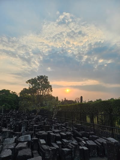 The image captures a serene outdoor scene at sunset in Bokoharjo, Indonesia, likely within the vicinity of Ratu Boko temple complex. The sky is a canvas of soft colors, with a golden sun setting low on the horizon, peeking through a layer of scattered, wispy clouds. The foreground is dominated by a collection of roughly cut, dark-colored stone blocks, artfully arranged as if remnants of an ancient structure. These stones are weathered and show signs of age. Beyond the stone blocks, a lush green landscape unfolds, featuring trees with dense foliage. A dark metal fence runs horizontally across the middle ground, separating the stone area from the greenery beyond. The overall impression is one of tranquility and historical resonance, with the natural beauty of the sunset complementing the ancient stone remnants.
