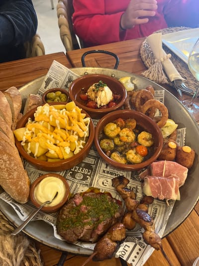 Here is a description of the image:
The image shows a high-angle view of a large, round metal tray filled with a variety of tapas-style dishes. The tray sits on a wooden table with a decorative woven mat visible beneath it. A child's hand clad in a red sleeve is partially visible at the upper left corner of the image, suggesting the food is being shared or enjoyed at a table.
The tray contains several small brown bowls and individual servings of food items arranged on newspaper-style paper with vintage advertisements and graphics printed on it. The dishes include:
* Fried potatoes drizzled with sauces.
* Garlic Shrimp.
* Burrata with tomatoes.
* Skewered meat.
* Croquettes.
* Prosciutto wrapped around mozzarella balls.
* A steak cut and topped with herbs.
* A bowl of mayonnaise with a spoon.
* A side dish of olives.
* A portion of bread.
* Onion rings
The composition of the tray suggests a shared meal or tasting experience at a restaurant, likely in Xàbia / Jávea, Spain, a popular tourist destination.