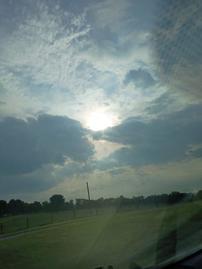This is a photograph taken from inside a vehicle, looking out at a landscape under a cloudy sky. The sun is partially obscured by clouds, casting rays of light. The foreground is a grassy field with a paved path running through it, bordered by a fence. In the background, there is a line of trees and a utility pole. The photograph appears to have been taken in Stamping Ground, United States, during daylight hours, possibly in the late afternoon or early evening given the angle of the sun and the cloud cover.