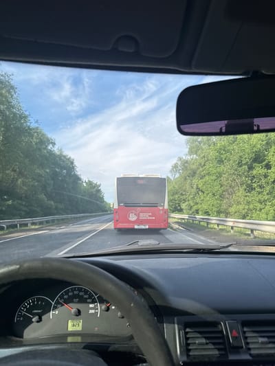The image presents a first-person view from inside a moving vehicle, looking forward through the windshield. The scene depicts a road in Усть-Сыны, Russia, on a bright, sunny day with a blue sky and scattered white clouds.

In the foreground, elements of the car's interior are visible. On the right, a portion of the steering wheel and the dashboard instrumentation are seen, including a speedometer showing a speed of approximately 40-50 km/h and an odometer reading 198906 km. A digital display indicates "1829", likely a trip meter. A red triangular hazard light button is also visible on the dashboard. On the left, the car's rearview mirror hangs, reflecting a subtle purple tint.

The primary subject outside the car is a red bus ahead, which the car is following. Its rear is clearly visible. The roadside is lined with dense green trees and a metal guardrail.

Visible text on the bus includes "ПЕРМСКИЙ", "ПЕРИОД", and "НОВОЕ ВРЕМЯ", which translates to "Permian Period New Time" or "Perm Region, New Time," indicating the bus's operational area is likely the Perm region of Russia. A vehicle number or license plate "1205ac1" is also present on the bus. The car's dashboard features standard numerical markings for speed (0-200 km/h) and engine RPM (x1000min).