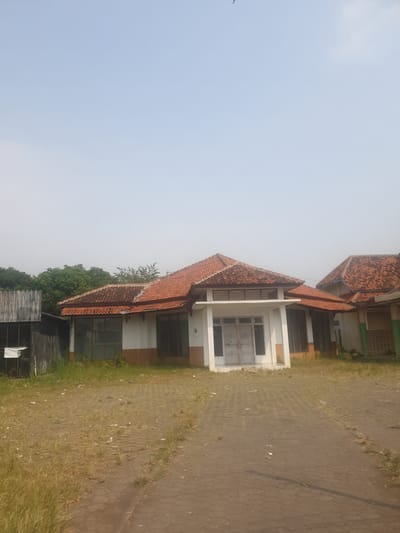 The image shows an abandoned building with a red tiled roof in Watesalit, Indonesia. The building appears to be a single-story structure with a prominent entrance featuring double doors. The exterior walls are painted white and brown. Large windows are visible on the front of the building. The surrounding area is overgrown with grass and weeds, suggesting a lack of maintenance. A paved walkway or driveway leads to the building. In the background, there are trees and what appears to be another section of the building, also with a red tiled roof. The sky is clear and bright, indicating daylight. There are no people or any apparent activity in the scene. The overall impression is one of neglect and disuse.