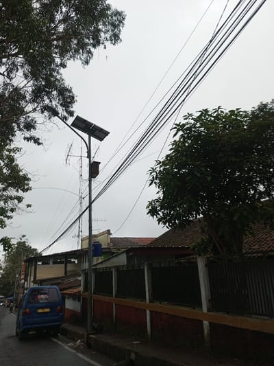 The image presents an overcast street scene in Ambakirano, Madagascar. The sky is uniformly light grey, occupying the upper half of the frame. In the foreground, a dark asphalt road extends from the lower left towards the mid-ground. A blue van is parked on the left side of the road, facing away; its yellow license plate displays partial characters. Adjacent to the road's right edge, a dark metal fence with vertical bars is situated atop a red and orange painted low wall. A gutter containing minor debris runs parallel to the wall.
A grey utility pole stands centrally, equipped with a solar panel tilted upwards and a street light fixture extending horizontally to the left. Below the solar panel, multiple communication antennas are attached, alongside a brown electrical box. Numerous black utility cables originate from this pole, traversing diagonally across the sky to the right and descending downwards.
Dense green foliage of a large tree is visible in the upper left quadrant. A smaller, bushy tree with dark green leaves is positioned on the right side, behind the fence. In the mid-ground, behind the utility pole and fence, various buildings with tiled roofs, some terracotta-colored, are discernible. One building, left of center, features a light-colored upper section and a blue cylindrical water tank mounted next to it. No individuals or specific actions are clearly depicted.