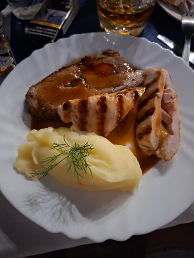The image presents an overhead view of a prepared meal served on a white, scalloped-edge plate. The plate holds a variety of food items, including three distinct portions of meat and a scoop of mashed potatoes. On the left, a dark cut of meat, possibly beef or pork, is covered in a rich brown gravy. Above it, several slices of grilled chicken are visible, featuring distinct char marks from the grill; some slices appear to be stuffed or layered with another ingredient. On the right side of the plate is a substantial serving of creamy, pale yellow mashed potatoes, garnished with a sprig of fresh dill.

The setting appears to be indoors, likely in the evening or at night, indicated by the dark background and prominent artificial lighting that creates reflections on the plate and food. To the left of the plate, a glass containing an amber-colored drink, possibly an alcoholic beverage like whiskey, is partially visible. A silver fork also peeks into the frame from the top left. In the bottom left corner, a Marlboro Gold cigarette pack is clearly visible, displaying its brand name and a health warning in what appears to be Bulgarian text. A colorful patterned lighter is positioned next to the cigarette pack. The scene depicts a casual meal, possibly dinner, in Ruska Bela, Bulgaria, as per the provided context.