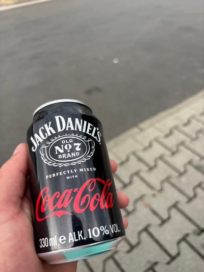A person's hand holds a black aluminum can outdoors, resting on a paved surface made of interlocking grey bricks. The can is labeled "JACK DANIEL'S OLD NO. 7 BRAND" and "Coca-Cola" in white and red respectively. Text below the Coca-Cola logo reads "PERFECTLY MIXED WITH" and "330 ml e ALK. 10% VOL.". The can appears to be a ready-to-drink alcoholic beverage. The lighting suggests it is daytime, and the background is blurred, but the visible texture of the paving stones indicates an outdoor, possibly urban or suburban setting. The focus is on the can and the hand holding it.