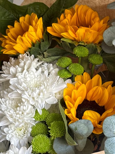 The image shows a close-up, overhead view of a floral arrangement. The arrangement features bright yellow sunflowers with dark centers, white chrysanthemums with many delicate petals, and small, round, green button-like flowers. There are also some muted green leaves and greyish-green eucalyptus leaves interspersed throughout the bouquet. The background is out of focus and appears to be a light brown, textured surface, possibly a table or a box. The lighting is bright and natural, highlighting the vibrant colors and textures of the flowers. There are no people, actions, or interactions visible in the image. The image is a still life of a flower arrangement. There is no information in the image that would allow for a determination of its location in the city of Novopokrovka, Kyrgyzstan.