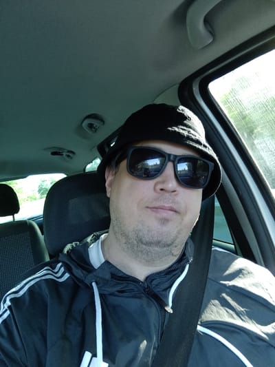 The image is a selfie taken by a man in the driver's seat of a car, appearing to be in motion. He is wearing a black bucket hat, black sunglasses, and a black hooded jacket with white stripes. The man has short, stubbly facial hair and is looking directly at the camera. The interior of the car is visible, including the dashboard, ceiling, and part of the passenger seat. Sunlight streams in from the right side, illuminating the scene and casting shadows. The reflection in the sunglasses shows a road and trees, suggesting a rural or suburban setting. There is no specific indication of the location being Mladá Boleslav, Czechia.