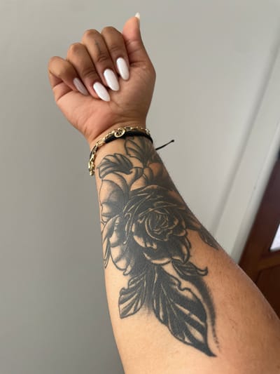 The image presents a close-up, slightly overhead view of a person's left forearm and hand, highlighting a significant tattoo and manicured nails. The central subject is a large, intricate blackwork tattoo that covers the entire forearm. The tattoo design features multiple detailed roses and intertwining leaves, rendered in solid black and varying shades of grey.

The skin tone of the arm is tan, with fine hair visible on the upper arm. A delicate gold chain bracelet is worn on the wrist, intertwined with a black cord that includes a small circular charm. The hand is gently curled, with the fingers slightly bent. The fingernails are long, almond-shaped, and finished with a glossy, opaque white polish. A faint, light-colored mark or tattoo is barely discernible on the side of the index finger.

The background is a plain, light gray or off-white surface, likely a wall, which provides a clean and contrasting backdrop for the arm. A darker, possibly wooden, architectural element is visible in the top right corner. The lighting is bright and even, suggesting an indoor setting with natural or well-distributed artificial light. The image captures a static pose, showcasing the details of the arm and its adornments. No text is visible within the image. The location context for this image is Krainitsi, Bulgaria, though no specific visual cues for this location are present in the frame.