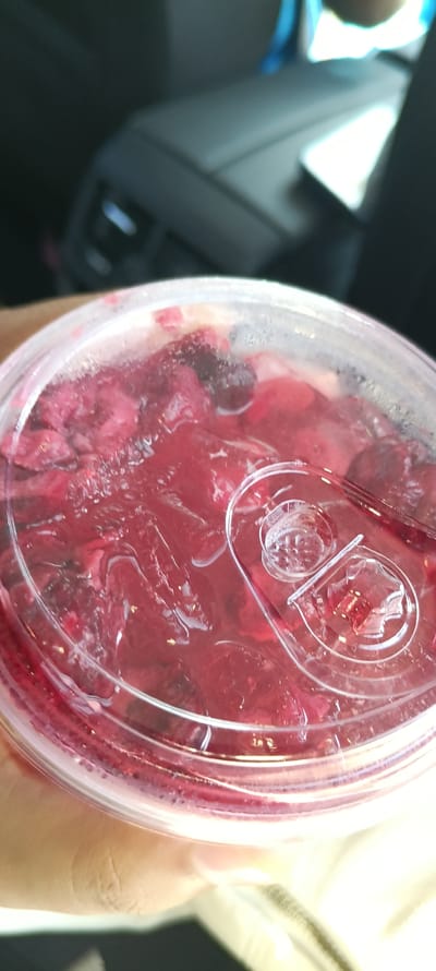 A close-up, overhead shot shows a clear plastic cup filled with a dark red, icy beverage, possibly a fruit drink with berries. The cup has a clear plastic lid with a built-in straw holder, and condensation can be seen on the outside of the cup. The beverage contains visible pieces of what appear to be raspberries or cranberries. In the background, the blurred interior of a car is visible, including a headrest and part of the dashboard. The lighting suggests it is daytime. The image was taken in České Budějovice, Czechia.