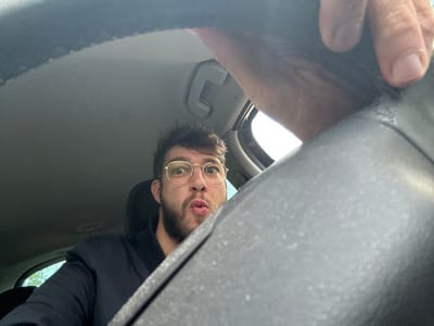 A low-angle photograph taken inside a vehicle in Scherwiller, France, depicts a single male individual. The male, positioned in what appears to be the driver's seat, has light skin, a visible beard, and wears gold-rimmed circular eyeglasses with a dark blue bomber-style jacket. His eyes are wide open, and his lips are pursed forward. The upper right portion of the frame shows a hand gripping a multi-textured, dark gray steering wheel, which partially obscures the lower right view. The car's interior ceiling is light gray, and a side window on the left reveals ambient daylight conditions outside. No other persons or distinctive external landmarks are identifiable.