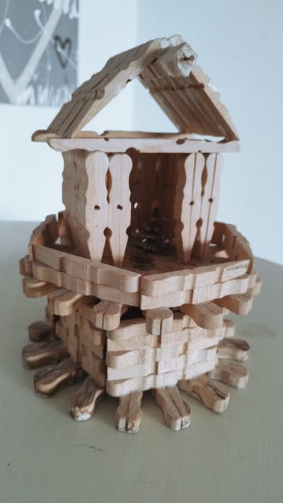 This is a medium-shot, eye-level, still photograph of a handmade wooden model of a house. The model is constructed from what appear to be clothespins and other small pieces of wood. The house has a multi-tiered structure with a pitched roof. The lower section is made of stacked wooden pieces arranged horizontally and vertically, with outward-facing wooden pieces forming a decorative base. The middle section also features horizontal wooden pieces, and it is supported by vertical wooden elements that resemble clothespins, creating an open, hollow interior. Within this interior, a small pinecone is visible, suggesting a miniature natural scene. The upper section consists of vertical wooden pieces that form a structure beneath a gabled roof, also made of wooden slats. The entire model is made of light-colored wood, with some darker wood grain visible.
The model is placed on a light-colored, flat surface, likely a table, with a plain, light-colored wall in the background. To the left in the background, a portion of a framed artwork is visible, featuring abstract lines and a stylized heart shape in shades of gray and white. The lighting appears to be natural and soft, casting subtle shadows. There is no indication of time of day, weather, or specific emotions. No text is visible in the image. The context of Cherbourg-en-Cotentin, France, is provided but not visually discernible from the image itself.