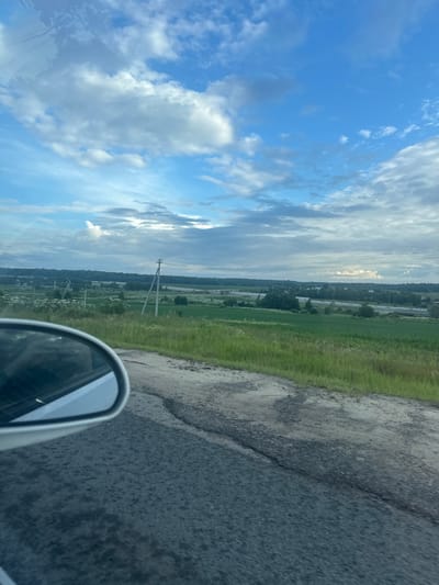 The image is a view from a moving vehicle, looking out over a rural landscape under a cloudy sky. In the foreground, a portion of a white car's side mirror is visible on the left, reflecting a blurred image of the car's interior. The road surface in the foreground appears worn and cracked. Beyond the road, a green field stretches out, dotted with white wildflowers. Several utility poles with wires traverse the field. In the distance, a line of trees forms a darker horizon, with a hint of buildings and infrastructure, possibly a highway overpass, suggesting the proximity of a more developed area. The sky is a mix of blue and white clouds, with patches of sunlight breaking through. The overall impression is that of a scenic drive through the countryside near Vvedenskoe, Russia.