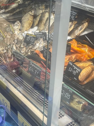 The image shows a display case of dried and smoked fish in a store. Several price tags with handwritten prices and names of fish are visible. The price tags are in Cyrillic script. The fish are arranged in trays and on shelves. The overall setting appears to be a food market or grocery store. Based on the context provided, this scene is located in the city of Гожули, Ukraine.