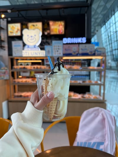 A person's hand, adorned with a polka-dot painted nail, holds a clear plastic cup filled with soft-serve ice cream and waffle cones. The ice cream is topped with chocolate sauce. In the background, a blurred retail environment is visible, featuring shelves stocked with various food items and a sign that reads "DRINKS." Further back, a white bear-shaped mascot is displayed. The scene appears to be indoors, possibly in a cafe or food establishment. The general atmosphere is casual and commercial, with the soft lighting and retail displays suggesting a public space.