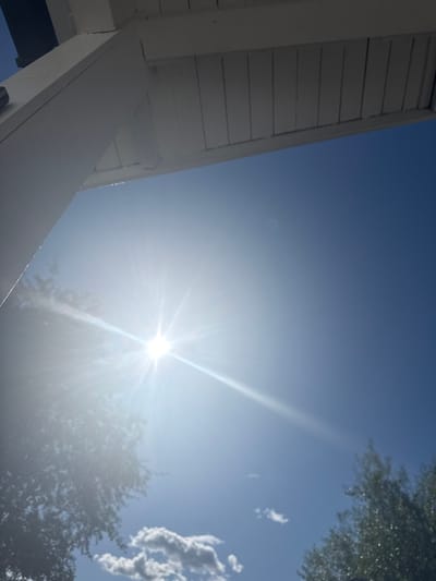 This is a low-angle, wide shot of a bright sunny sky taken from under a white wooden overhang, possibly a porch or awning. The sun is prominently featured in the center-left of the frame, emitting bright rays and lens flare. The sky is a clear, deep blue, with a few wispy clouds visible lower down. The edge of the wooden structure, with its slatted ceiling and white support beams, occupies the top portion of the image. There are hints of trees with green foliage at the bottom left and right edges of the frame, suggesting an outdoor setting. There are no people or specific landmarks visible to pinpoint a location within Moelv, Norway.