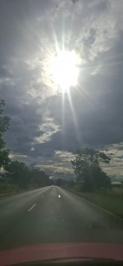 The image captures a perspective from within a vehicle, looking out at a paved road stretching into the distance. The sky above is dramatic, with the sun a bright, star-like burst at its zenith, partially obscured by dark, textured clouds that create a strong contrast. Rays of sunlight emanate from the sun, piercing through the clouds and casting beams downwards. The road itself is wet, reflecting the light and appearing somewhat slick. Lane markings are visible, indicating a two-lane road. Trees line both sides of the road, with some appearing lush and green, while others are darker and more dense. The overall atmosphere is moody and somewhat overcast, despite the bright sun. The location is specified as Gabarevo, Bulgaria. There are no people or discernible actions depicted, only the environment and the road.
