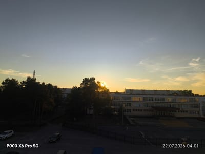 The image shows an outdoor scene at dawn in Kogalym, Russia. A school building is visible on the right, with the sun partially obscured by trees on the left, casting a warm glow. The sky is a gradient of blue and yellow, with wispy clouds. In the foreground, a paved area has a black metal fence. Two cars, a white hatchback and a dark sedan, are parked in the lower left corner. The timestamp "22.07.2026 04:01" is visible in the lower right corner, along with the text "POCO X6 Pro 5G" in the lower left. The overall impression is a quiet urban morning.