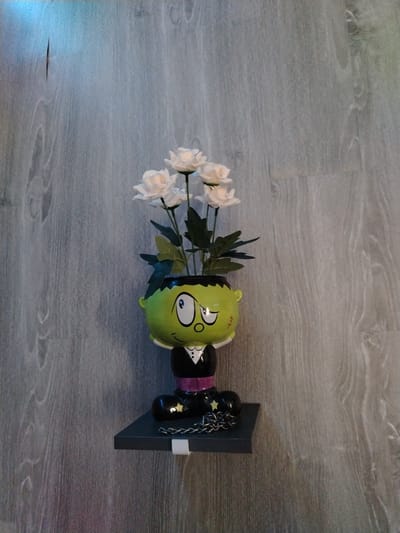 This media file features a novelty flower pot shaped like a cartoon character, possibly inspired by Frankenstein's monster, holding a bouquet of white roses. The pot is bright green with large eyes, stitch marks on the face, and a tuxedo-like outfit. It rests on a small, dark gray shelf mounted on a light gray wooden-paneled wall. The character's feet are adorned with black boots and a silver chain with star accents. The lighting appears to be artificial, casting subtle shadows and highlighting the textures of the wall and the flower pot. The overall scene is still and decorative, with no apparent activity taking place. The image is a close-up shot, focusing on the flower pot and the shelf against the backdrop of the wall. There is no visible text in the image. The location is identified as Vostochnaya, Russia.