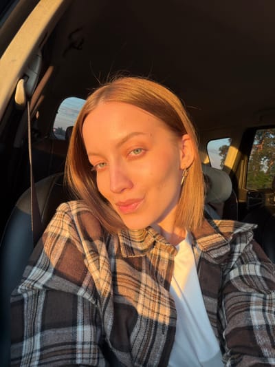 A young woman is photographed from a low angle while sitting inside a car. She is fair-skinned with light brown hair and green eyes. She is wearing a white t-shirt underneath a brown and tan plaid shirt. Golden sunlight streams into the car, illuminating her face and clothing. The interior of the car is visible, including the black leather seats and a window showing foliage outside, suggesting a natural or suburban environment. There is no discernible information about the location being in the city of Rogachovo, Russia.