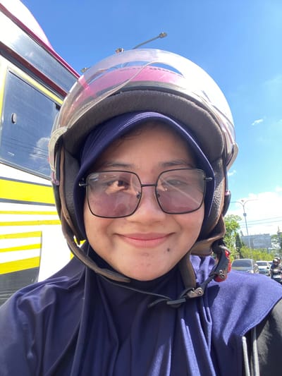 In Keraton, Indonesia, a young Indonesian woman wearing a blue hijab and sunglasses takes a selfie while riding a motorcycle. She is wearing a pink helmet, with the visor open. A large yellow and black bus is visible to her left. The sky is clear and blue with a few white clouds. Traffic, including cars and other motorcycles, is visible in the background.