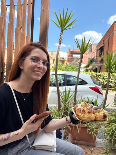 The image shows a woman with long red hair and glasses, smiling and holding a plate of cream puffs in her right hand. She is wearing a black top and a plaid jacket. In her left hand, she holds a smartphone. The background features a city street, a white car, and palm trees. The photo appears to have been taken outside, possibly at a cafe. Based on the presence of cream puffs specific to the Tivoli, Lazio region, the image is likely taken in Tivoli, Italy.
