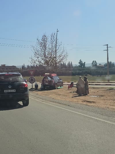 The image depicts an outdoor scene in Boufakrane, Morocco. A paved road runs across the lower third of the frame, with a dirt shoulder bordering its right side. On the road in the left foreground, a dark Volkswagen Touareg SUV, bearing Moroccan license plate "62314 | I 8", is partially visible, facing away from the viewer. Behind this vehicle, a red triangular "Give Way" sign and a circular "Mandatory Roundabout" sign are positioned.

On the dirt shoulder to the right, a dark SUV with a rear-mounted spare tire and a roof rack carrying various items is parked, facing right. Two red folding chairs are placed on the dirt shoulder between the parked SUV and two individuals. These two individuals, dressed in light-colored traditional garments that cover their bodies and heads, are standing and appear to be interacting.

In the midground, behind the parked SUV, a deciduous tree with sparse leaves stands adjacent to a lamppost. Power lines traverse horizontally across the upper midground, supported by a utility pole on the right side. The background features an extensive area of green fields or orchards populated by numerous trees. Distant buildings are visible on the far left horizon, and subtle geographical elevations are discernible on the far right horizon under a clear blue sky.
