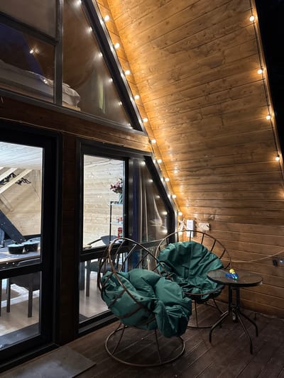 The image depicts an outdoor balcony area of a wooden A-frame structure, likely a guesthouse or cabin, at night. The space is illuminated by string lights draped along the wooden walls and a large triangular window. Two large, round, metal chairs with plush green cushions are positioned on the wooden deck. A small, round black table with a glass top stands between the chairs, holding a small blue and yellow toy water gun and a bottle. Through the large glass doors, another interior space is visible, featuring wooden walls and ceiling, and several chairs and tables, along with what appears to be kitchenware. The overall ambiance is cozy and rustic, with a view of what seems to be an interior loft space with a bed visible through the triangular window. The scene is devoid of people. Given the context provided that this is in Arkhyz, Russia, the environment suggests a mountainous or rural setting often associated with ski resorts or nature retreats.