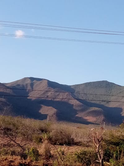 The image displays an arid, mountainous landscape located in La Cruz del Pastel, Venezuela, under a clear blue sky. In the upper portion, a single small white cloud is present in the upper-left quadrant. Four parallel power or communication lines traverse the upper-middle section of the frame horizontally. The mid-ground is dominated by a range of rugged, sparsely vegetated mountains, characterized by undulating peaks and valleys, with distinct shadows indicating varied topography and direct sunlight. The predominant colors of the mountains are shades of brown, dark green, and grey. Visible within the mountainous terrain are at least two lattice-structured power transmission towers. The foreground consists of dry, scrubby terrain with sparse, desiccated vegetation, including numerous bare branches and some low-lying green foliage. No individuals or discernible animal life are present. The scene is illuminated by natural light, casting pronounced shadows.