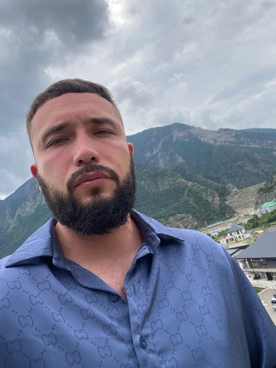 A man with a beard and short hair, wearing a blue shirt with a repeating "GC" pattern, stands outdoors against a backdrop of mountains. The sky is overcast with clouds. In the distance, several buildings and infrastructure are visible along a hillside, suggesting a partially developed or mountainous urban area. The overall setting appears to be in a mountainous region, potentially near Chalda, Russia, given the architectural styles and terrain. The man is looking towards the camera, and the image is a close-up of his upper body and face.