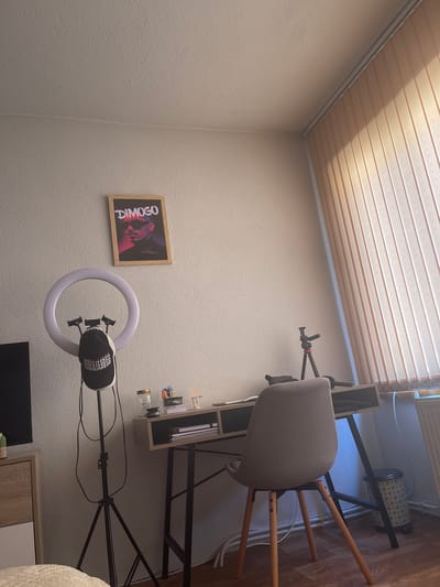 The image shows an indoor scene, likely a room, with a desk and chair as the central focus. A light-colored, textured wall dominates the left side of the frame, with a framed picture hanging on it. The picture displays a person's face with the text "DIMOGO" in large, white letters. To the right of the picture, a ring light with a black attachment sits on a tripod. The background reveals a wooden floor and a portion of a desk with shelves, where various items like pens and jars are visible. A grey chair with wooden legs is positioned behind the desk. The ceiling features horizontal wooden planks, suggesting it's a modern or renovated space. The lighting in the room appears to be a mix of natural light filtering through blinds from a window and artificial light from the ring light. The overall impression is that of a personal workspace, possibly set up for content creation or online activities. The scene is static, with no apparent activity occurring. The image is taken with the camera oriented in landscape mode, rotated counter-clockwise.