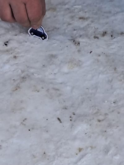 In Bolshaya Elhovka, Russia, a human hand is observed in the upper left quadrant, manipulating a small, flat, cutout representation of a dark blue compact car. The car object, outlined in white with visible white circular elements resembling wheels or lights, is positioned against a textured surface of dirty, partially melted snow. This ground surface is predominantly white with irregular light brown patches and scattered dark specks of debris. The hand appears to be placing or adjusting the car object on the snowy terrain.