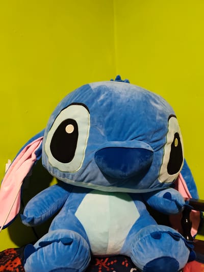 A close-up, eye-level shot shows a very large, plush toy Stitch character. The toy is predominantly blue, with large, oval eyes that have black centers and white pupils. Stitch's ears are long and pink on the inside, with a blue exterior. The toy's belly is a lighter blue. It is sitting on a surface with a dark, patterned fabric. The background is a solid, vibrant lime green wall, with the corner visible on the right. A thin white wire is partially visible on the left side of the toy, extending from behind it. The lighting appears to be artificial, casting some shadows on the toy and the background. The overall impression is of a brightly colored children's toy. The location is the city of Stryków, Poland.