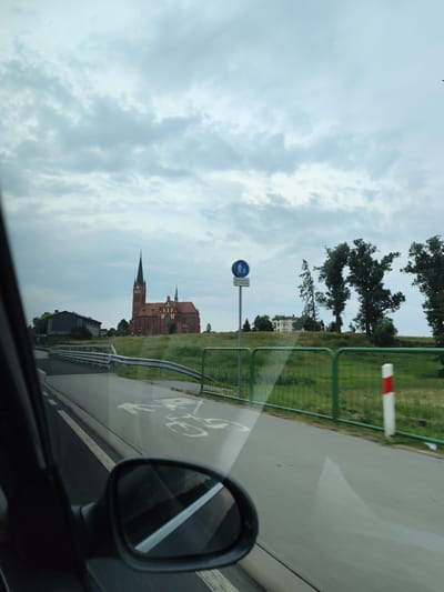 In Brzeźce, Poland, on a cloudy day, a view from a car shows a road with a bicycle lane marked on it. A guardrail lines the right side of the road, beyond which is a grassy area with trees. A red brick church with a tall spire is prominently visible on a hill in the background, along with a smaller building to its left. A street sign indicating a pedestrian crossing is also present. The car's side mirror reflects the road and sky.
