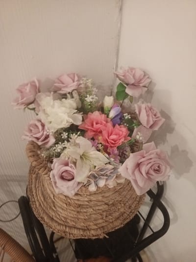 A floral arrangement is displayed in a woven basket. The arrangement features a variety of flowers, including pink roses, white flowers, and pink lilies. The basket is made of a natural, fibrous material and has a rustic texture. It sits atop a dark metal stand, which is partially visible. The background consists of a white wall with a subtle vertical texture. The lighting is soft, casting gentle shadows. The scene is devoid of any human presence.