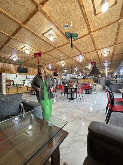 The image shows the interior of a cafe or restaurant. A woman is standing in the foreground, wearing a green dress and an olive green jacket. She is holding a pair of glasses in her right hand. The cafe has a wooden ceiling with exposed beams and decorative bows. There are tables and chairs, a counter area with food displayed, and a large fireplace in the background. The floor is tiled with a marble pattern. The cafe appears to be decorated for a holiday, possibly Christmas, with the red and green bows. The location is Tsovagyugh, Armenia.