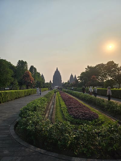 The image captures a wide-angle view of the Prambanan Temple complex in Tlogo, Indonesia, at sunset. The sky is a hazy orange and yellow due to the setting sun, which is visible in the upper right corner. A paved pathway, lined with meticulously manicured hedges and colorful flowerbeds, leads towards the towering central temple and several smaller spires in the distance.
Several people are seen walking along the pathway and on the grounds. Some are dressed in traditional Indonesian attire, including headscarves. They appear to be tourists or visitors exploring the historical site. The architecture of the temples is distinctively Hindu, with pointed spires characteristic of Javanese religious structures. The surrounding environment includes lush green trees and well-maintained gardens, creating a serene and picturesque atmosphere. The light suggests it is either sunrise or sunset, with a warm, diffused glow illuminating the scene.