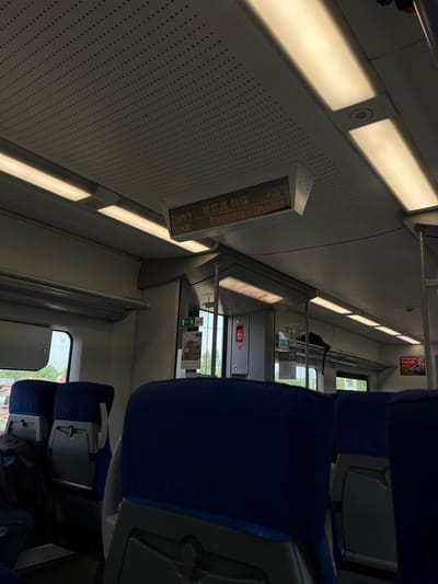 Here is a description of the image:
The image shows the interior of a train car. The ceiling is white with evenly spaced light fixtures. A display shows the date, time, temperature (26°C), and the station names "Alabushevo - Kryukovo". The speed is shown as 24 km/h. The seats are upholstered in blue. There is a window to the outside showing foliage. A sign with "100 рублей" is also visible. Based on the stations named on the display, this train is likely in or near Povarovo, Russia, as Alabushevo and Kryukovo are neighborhoods in the area of Zelenograd and located in the Moscow region.