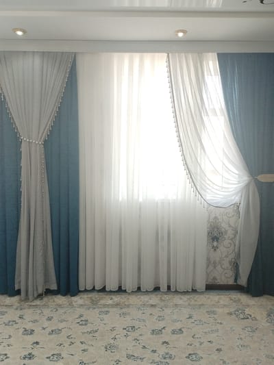 This is a full-shot, eye-level, indoor photograph of a window adorned with multiple layers of curtains and a patterned carpet covering the floor. The scene appears to be a room in a residential building located in Чагыр, Kyrgyzstan.

The window is draped with three sets of curtains. The outermost layer consists of two teal-colored, opaque curtains, one on each side of the window. The left teal curtain is partially drawn back, revealing a light gray sheer curtain layered beneath it. This gray curtain is held back by a decorative tie-back, a strand of pearls and beads that runs from the top of the curtain down to about its mid-point, where it is secured by a band of larger pearls. The gray curtain itself is adorned with multiple strands of smaller pearls and beads hanging vertically from its edges.

In the center, behind the gray curtain, is a white, sheer curtain that covers the entire window. This white curtain is the brightest element in the image, allowing a significant amount of light to filter through, obscuring the view outside. The white curtain is also decorated with strands of pearls and beads along its edges, and it is tied back on the right side with a light-colored fabric tie. The bottom edge of the white curtain is visible resting on the carpet.

The wall behind the sheer curtains has a faded, intricate wallpaper pattern in shades of gray and blue, featuring floral or damask motifs.

The floor is covered with a large, ornate carpet. The carpet has a light cream-colored base with a repeating pattern of blue floral designs and decorative borders.

The ceiling is white and features two recessed LED spotlights, one visible on the left side of the image and another on the right.

There are no people or animals present in the image. The overall impression is one of a well-decorated interior space.