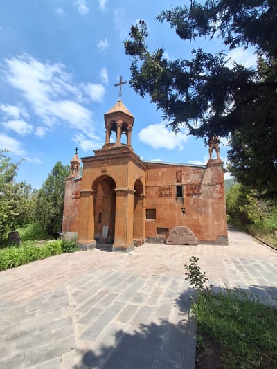 The image shows a red stone church with a bell tower, located in Martiros, Armenia. The church has an arched entrance and is decorated with stone carvings. There is a cross on top of the bell tower. The church is surrounded by trees and greenery, and the ground in front of it is paved with stone tiles. The sky is blue with some white clouds. A single, small tree with green leaves is growing in the paved area.