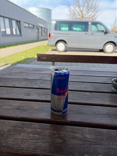 The scene depicts an outdoor break area in Bad Rappenau, Germany, under a clear sky. In the immediate foreground, a weathered dark brown wooden slatted picnic table occupies the majority of the lower frame. Centrally positioned on this table is an open, partially consumed Red Bull energy drink can, blue and silver, with its pull tab lifted. To the far right, a clear glass ashtray containing multiple cigarette butts and ash is partially visible.
Behind the table, a dark brown wooden bench runs horizontally. Beyond the bench, a paved light-colored surface serves as a parking area. Parked on this surface, facing right, is a light grey Volkswagen Transporter van, its side profile visible. To the left of the paved area, a strip of green grass separates it from a low, light grey industrial or commercial building featuring multiple rectangular windows with dark frames. Behind this building, a large, cylindrical, light-colored storage tank is visible. To the right, behind the van, a deciduous tree with bare branches stands against the horizon. The ambient light indicates a sunny daytime environment with discernible shadows. No individuals are present.