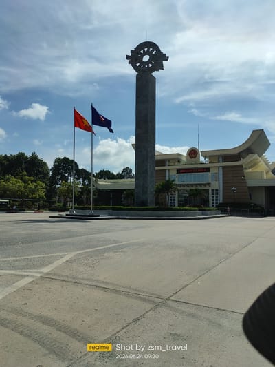 This is a wide-angle, eye-level shot of the Moc Bai border crossing in Ben Cau, Vietnam. In the foreground, a wide, paved area with white painted lines leads towards a large, stone monument topped with a wheel-like emblem. To the left of the monument are two flags: one red with a yellow star (Vietnam) and another dark blue with a gold emblem (ASEAN). Behind the monument and flags is a modern, multi-story building with a curved roofline. The building has large windows and signage that reads "CỘNG HÒA XÃ HỘI CHỦ NGHĨA VIỆT NAM" and "KÍNH CHÀO QUÝ KHÁCH" (Welcome guests). Several palm trees are planted in front of the building. The sky is blue with scattered white clouds. The overall scene suggests an international border crossing with official buildings and prominent national symbols. A portion of a person's dark clothing is visible in the lower right corner of the frame, likely indicating the photographer's presence. The image also contains a watermark in the lower left corner indicating it was taken by "zsm_travel" on "2026.06.24 09:20".
