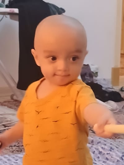 A young child with a shaved head, wearing an orange t-shirt with a bird pattern, is looking to the side and smiling. The child is holding a light-colored object in their right hand. The background is blurry and appears to be an indoor setting with various items of clothing and furniture visible. There is no information in the image that allows for the identification of the location as Yzgant, Turkmenistan.