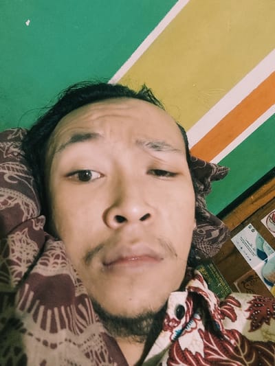 The image shows a close-up portrait of a man with dark hair. He is looking directly at the camera with a neutral expression. The man is wearing a red and white patterned shirt with a brown and white patterned fabric behind him. The image was taken in an indoor environment with a yellow, orange, and green striped wall in the background. The location based on the provided coordinates is Kecamatan Peterongan, Indonesia. The man’s face is mostly visible in the frame. He is not interacting with anyone. The image was taken with a handheld device at close range.