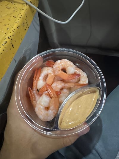 A person's hand, displaying a light to medium skin tone, holds a clear plastic container with two compartments. The larger compartment contains approximately ten to fifteen cooked, peeled, pinkish-orange shrimp. The smaller, oval-shaped compartment holds a quantity of light orange, creamy sauce with visible dark specks. The hand grips the container from underneath, with the thumb visible on the left side and fingers supporting the right. In the upper left background, a bright yellow object with multiple small black surface imperfections is partially visible. A white cable runs horizontally across the upper left of the frame, partially crossing the yellow object. The remaining background consists of dark, indistinct gray and black tones, with a blurred section of light blue-gray fabric visible in the lower right, suggesting an interior environment such as a vehicle or confined space. No specific visual information within the image indicates a precise location within Ungheni, Romania.